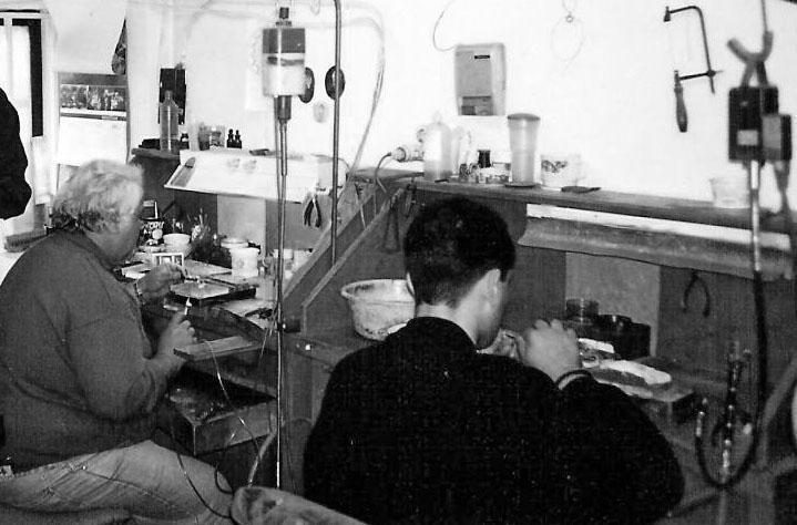 Two craftsmen at work in the Dimos workshop, photographed in black and white — bench tools, vintage equipment, hands focused on the work.