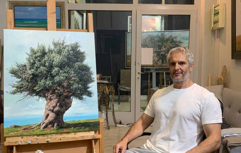 Elidon in his studio, seated beside one of his olive-tree paintings on an easel.