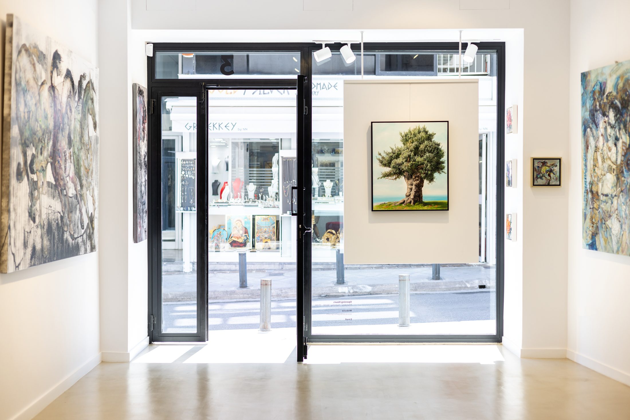 Inside Orlik Gallery: paintings on the walls, Elidon's olive tree framed in the centre, and the gallery doors open onto Plaka with the Dimos jewellery display visible across the street.