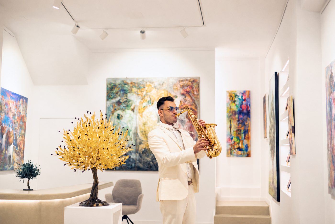 Tasos Sarioglou playing saxophone in a gallery, dressed in white, with abstract paintings on the walls and a sculpted gold-leaf tree on a plinth beside him.
